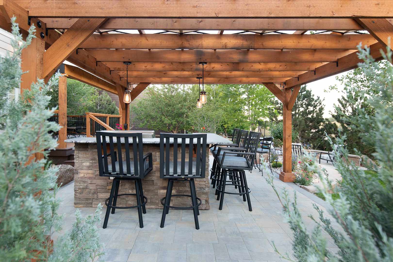 Artistic view from between two juniper trees of a modern pergola with built in light and a stacked stone bar with modern black barstools. 