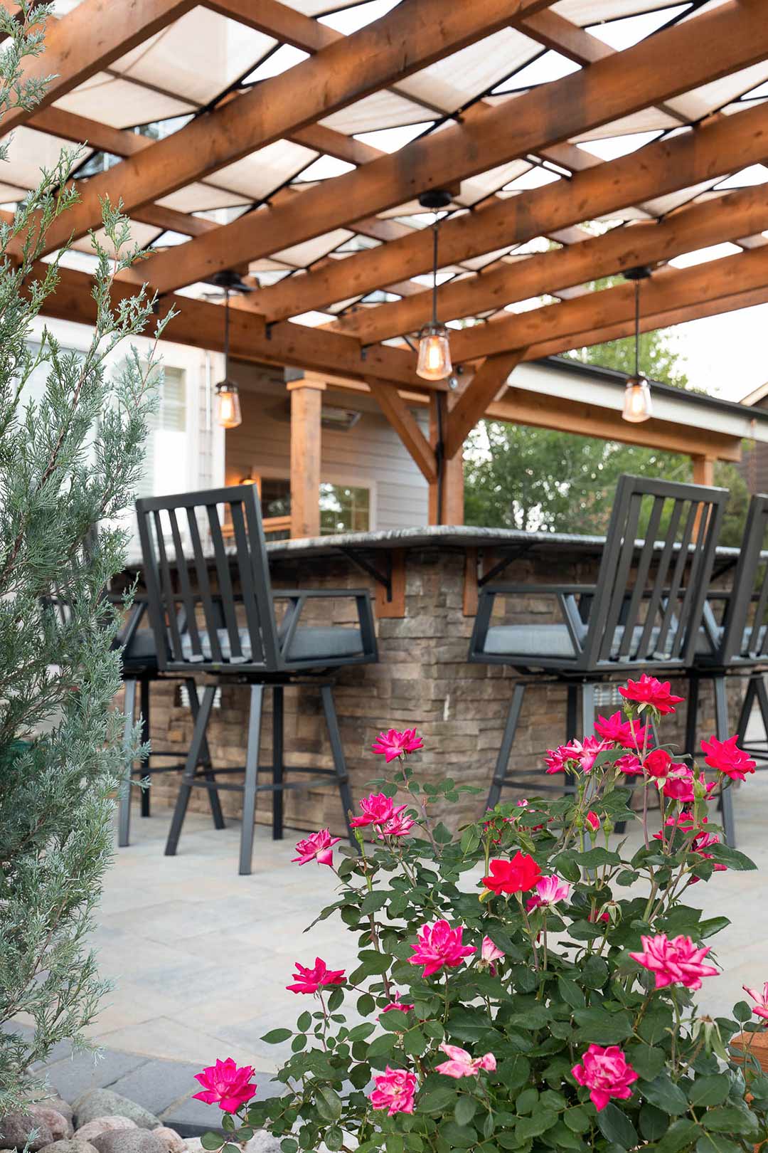 Artistic view of a modern pergola with built in light and a stacked stone bar with modern black barstools. 