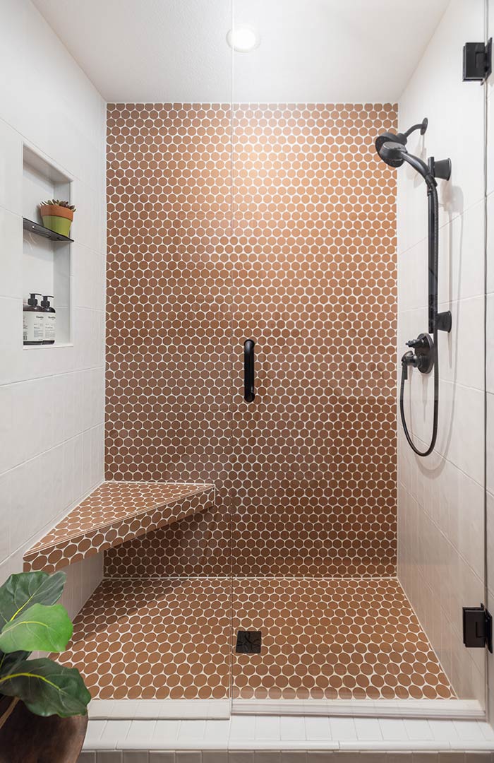 A beautiful walk-in shower with custom tile work with oversized copper penny tile and matte black hardware and bench created by Freestone Design-Build and their full service interior design package